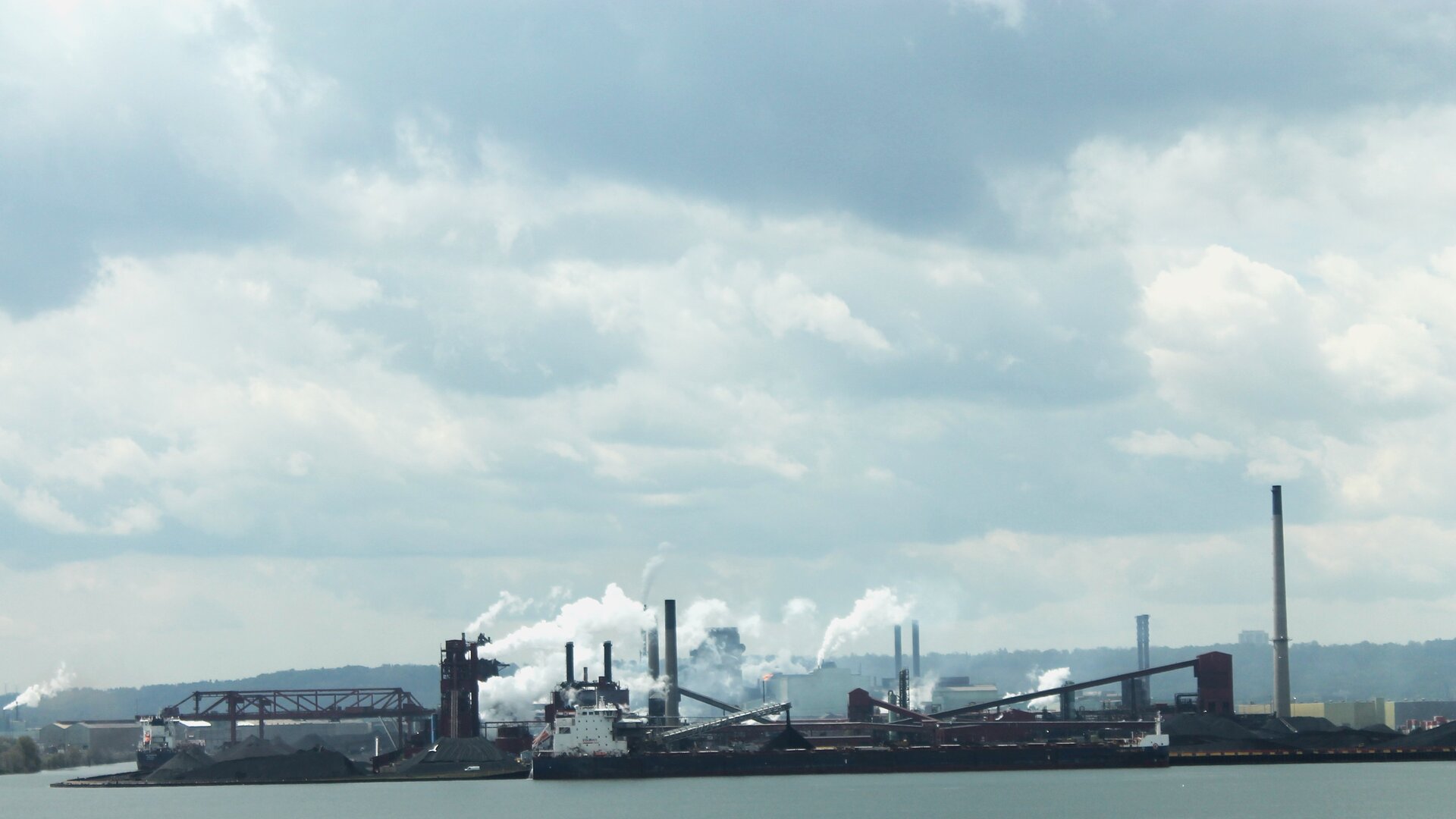 Hamilton steel mill seen from the waterfront, smoke rising from multiple stacks against a cloudy sky.