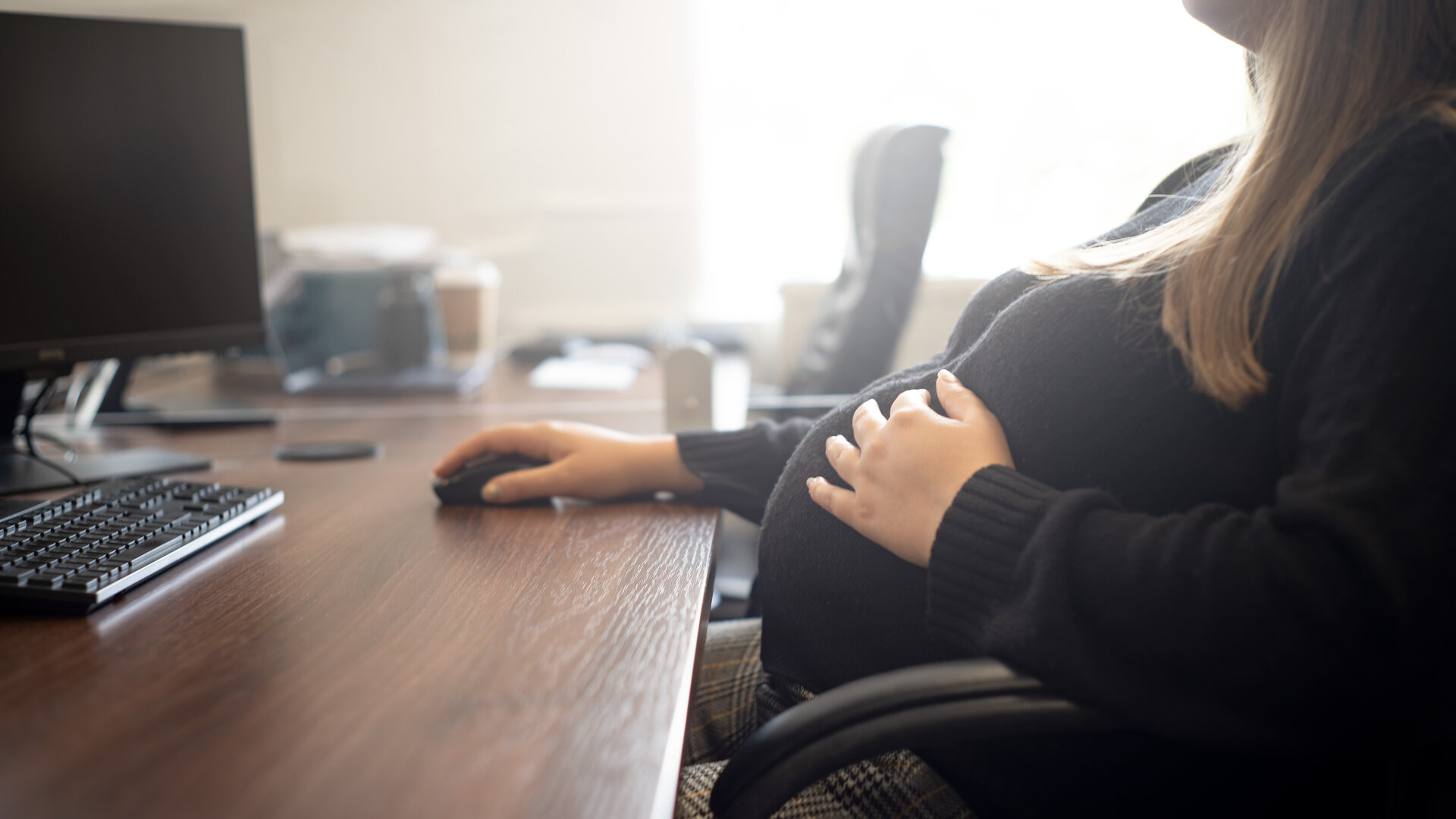 A pregnant woman at a wood-veneer office desk, hand resting on her belly, working at a computer.