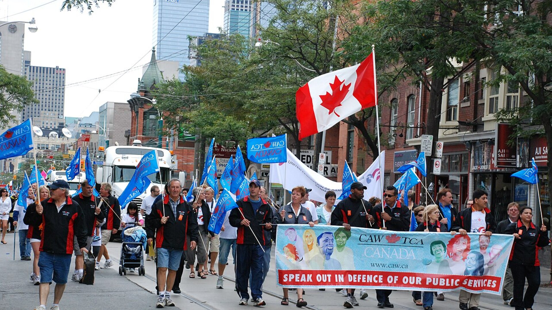 Union members marching in Toronto's 2011 Labour Day Parade behind a banner reading Speak Out in Defence of Our Public Services, with the downtown skyline behind them.